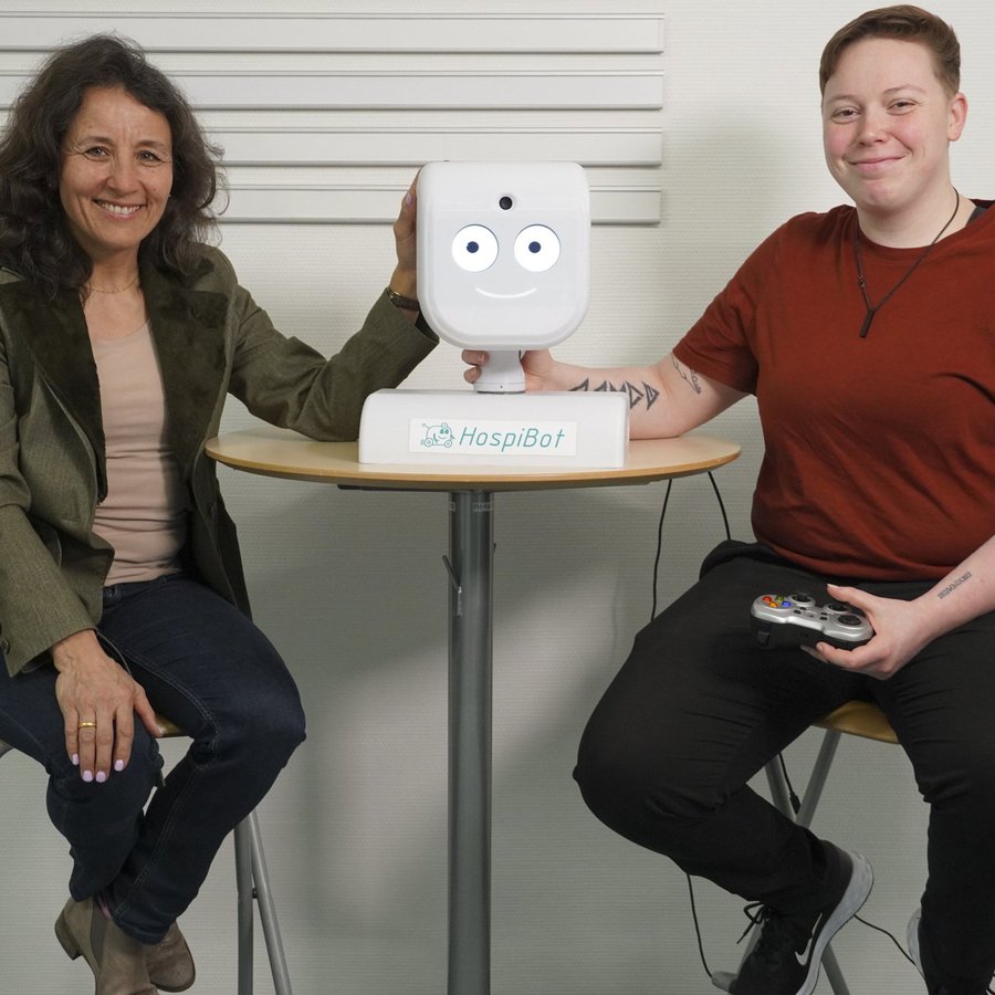 Zwei Frauen sitzen auf der rechten und linken Seite eines Bistrotischs, auf dem Tisch steht der Kopf eines Roboters.   