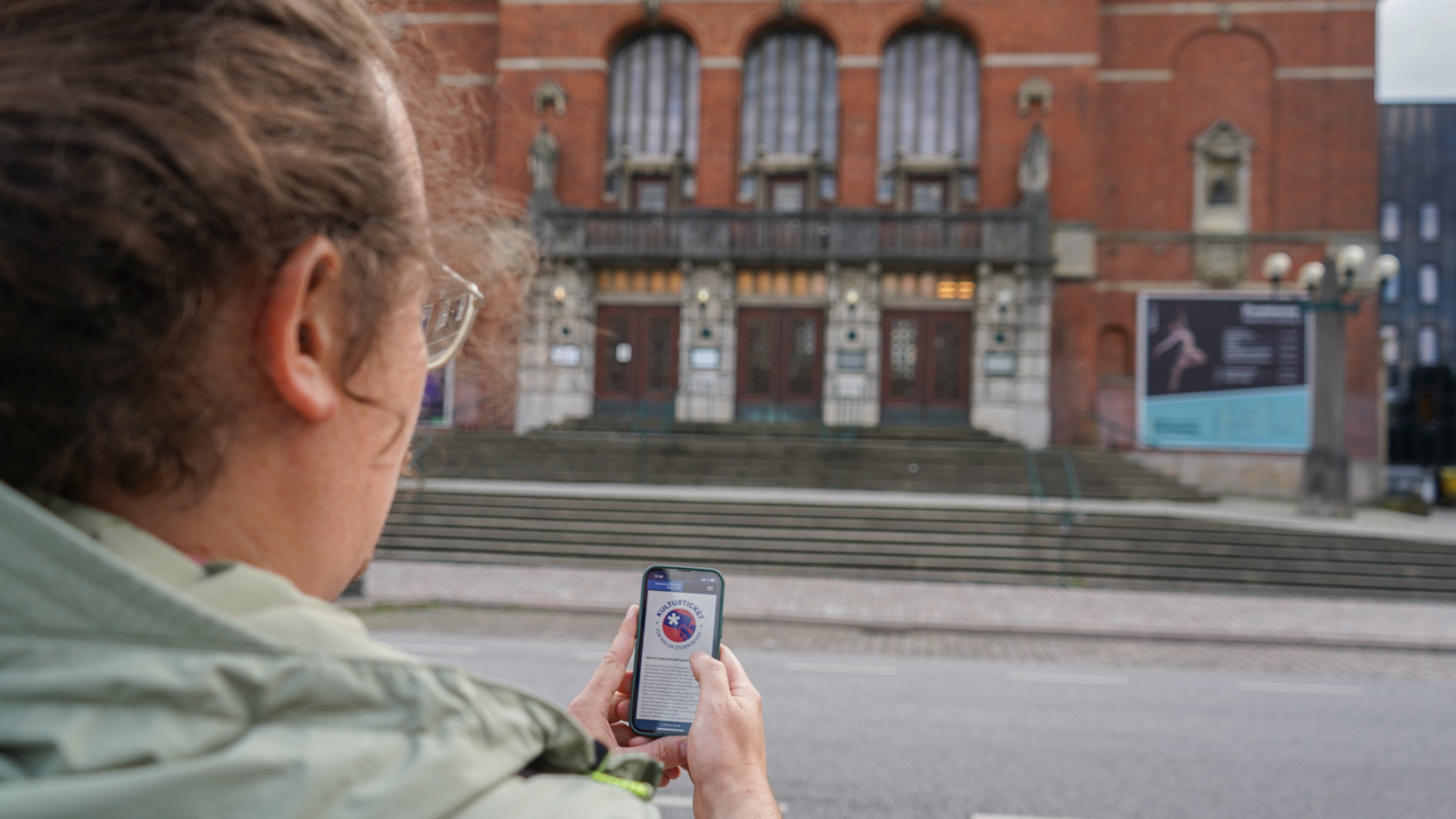 Ein Mann steht mit Handy in der Hand vor dem Kieler Opernhaus. Auf dem Handy sieht man die Website des Theaters, über die Studierendentickets gebucht werden können.