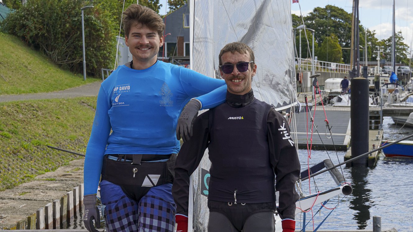 Zwei junge Männer in Segelkleidung stehen auf einem Steg.