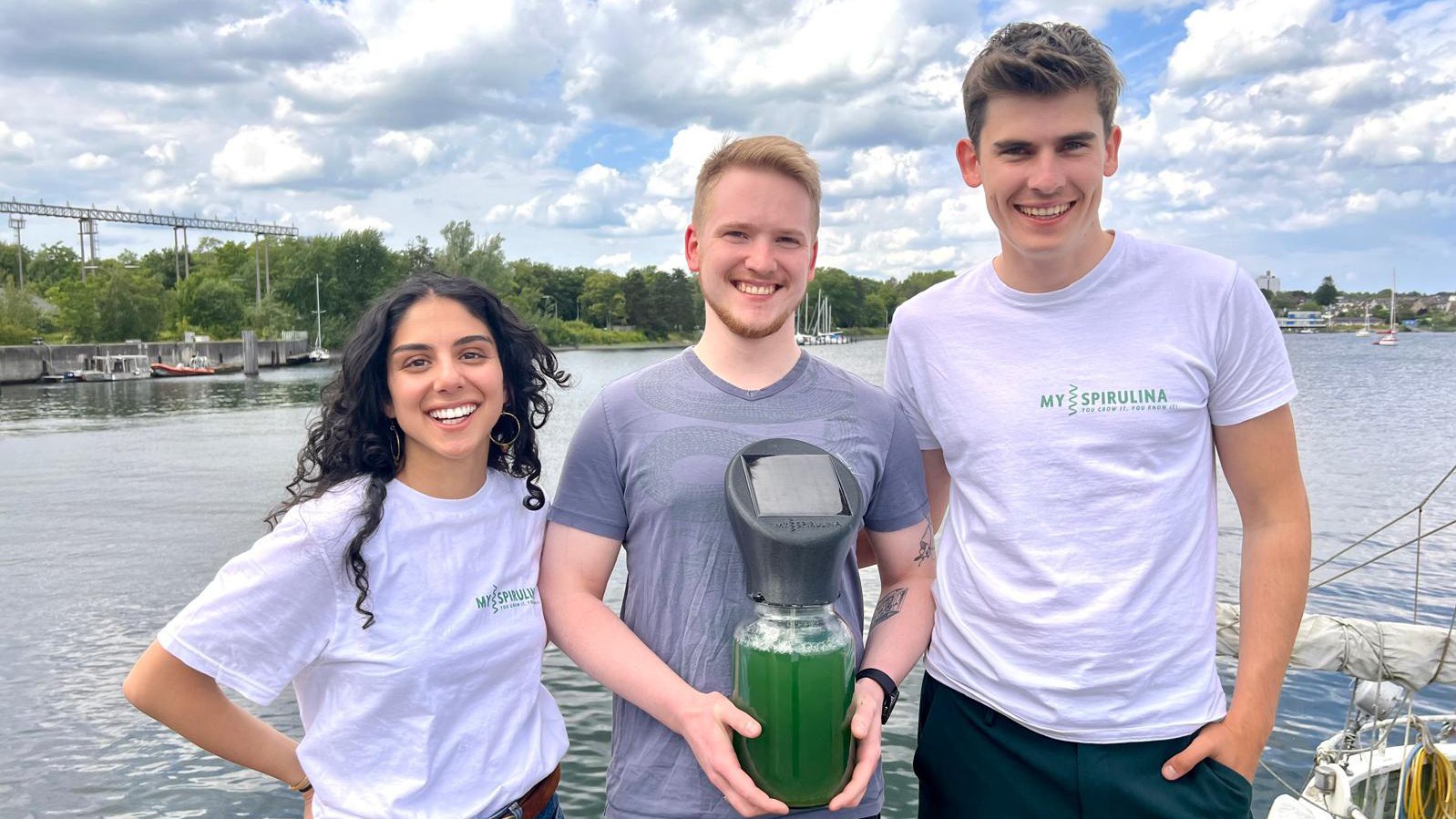 Die Gründer Sara Farzaneh Nejad, Jan Thode und Ben Schwedhelm stehen mit einem Glas Spirulina in der Hand am Waser..