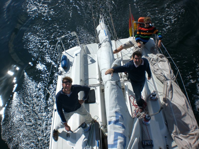 Auf dem Foto sieht man drei Männer, lässig auf einem Segelboot posieren.