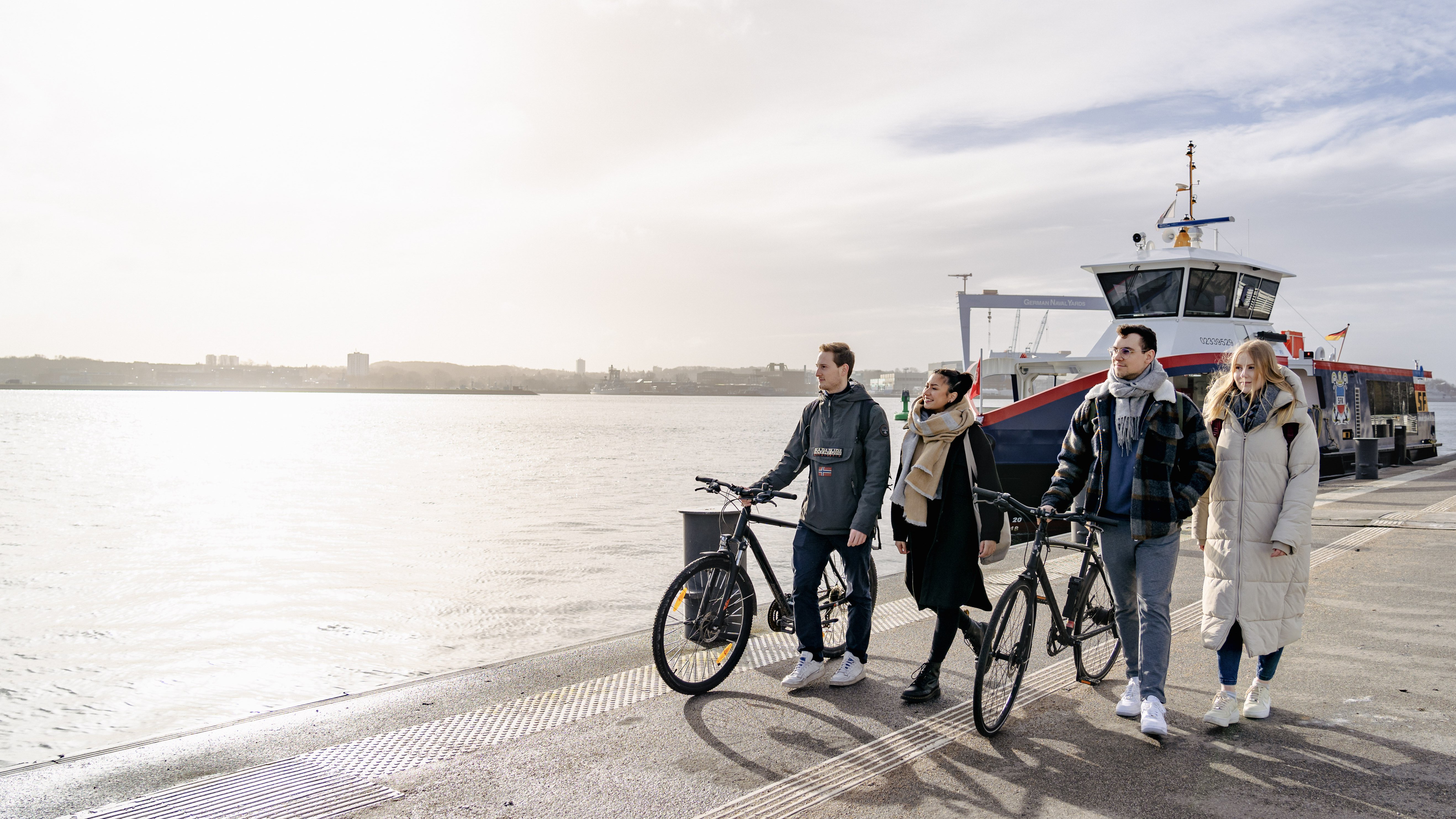 eine Gruppe von vier Personen geht entlang der Kiellinie im Sonnenschein, im Hintergrund ist der HDW-Kran zu sehen und eine Elektrofähre