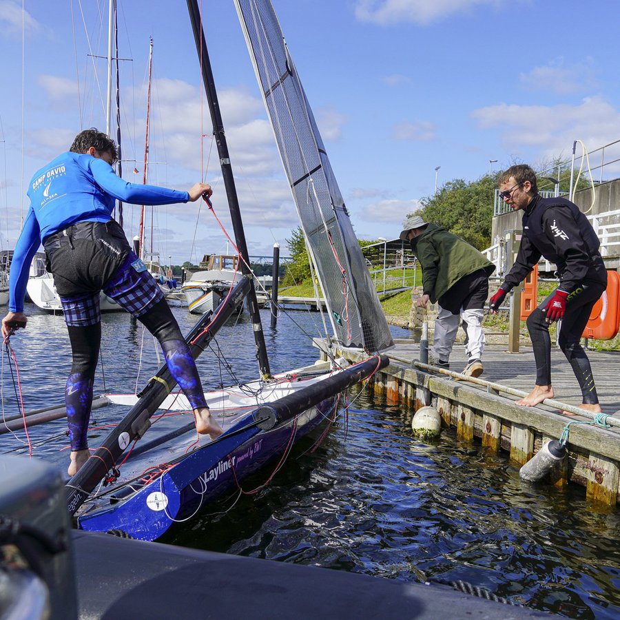 Zwei junge Männer stehen auf einem Holzsteg, einer auf einem Skiff, einem sportlichen Segelboot. 