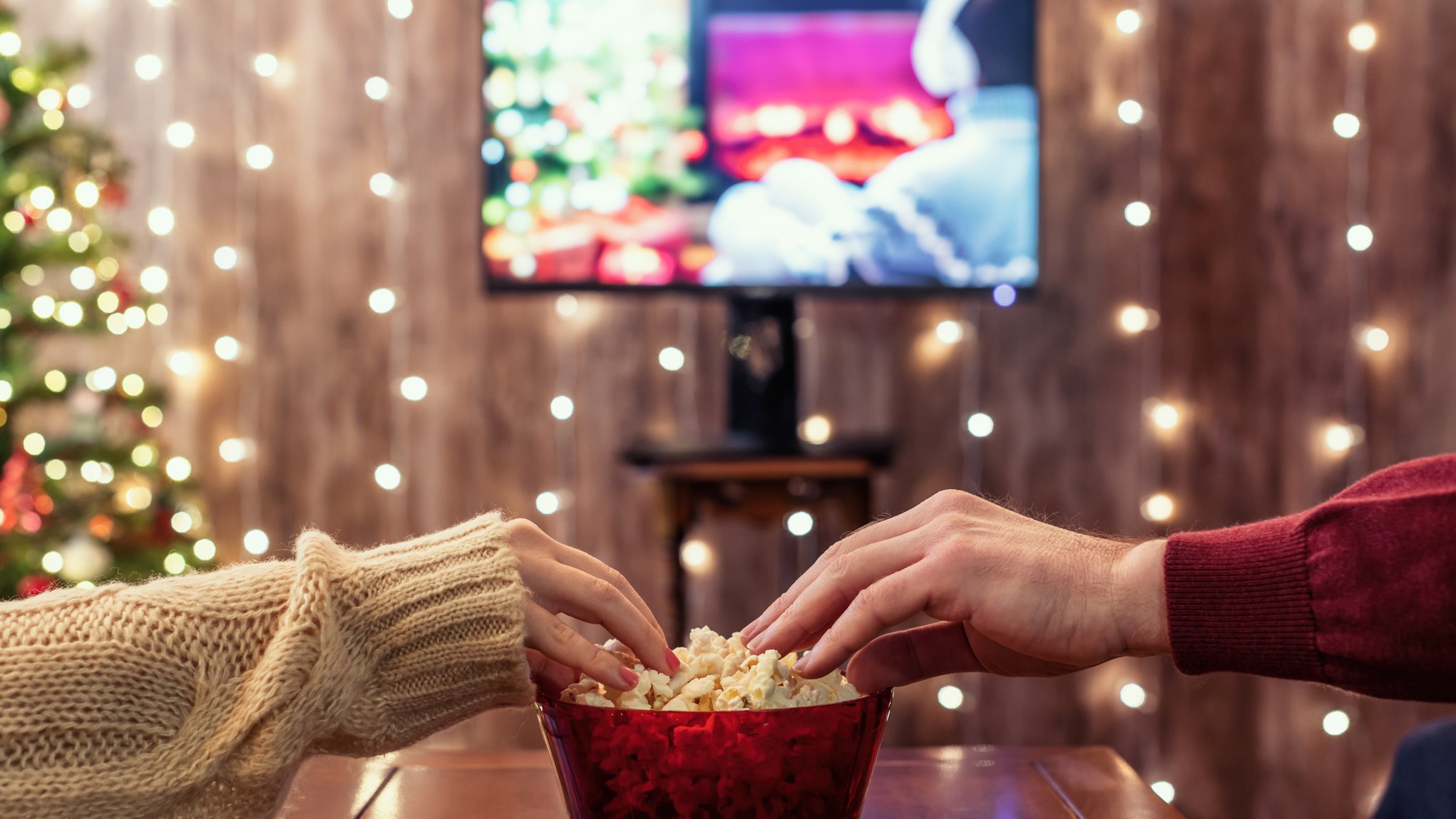 zwei Hände greifen in eine Popcornschale, dahinter ein Fernseher, drumherum weihnachtliche Dekoration