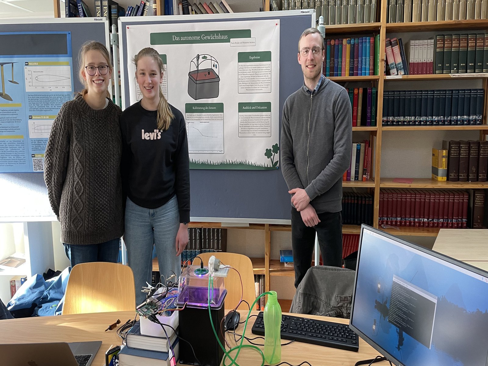 Zwei Schülerinnen und ein Student stehen vor einer Plakatwand. 