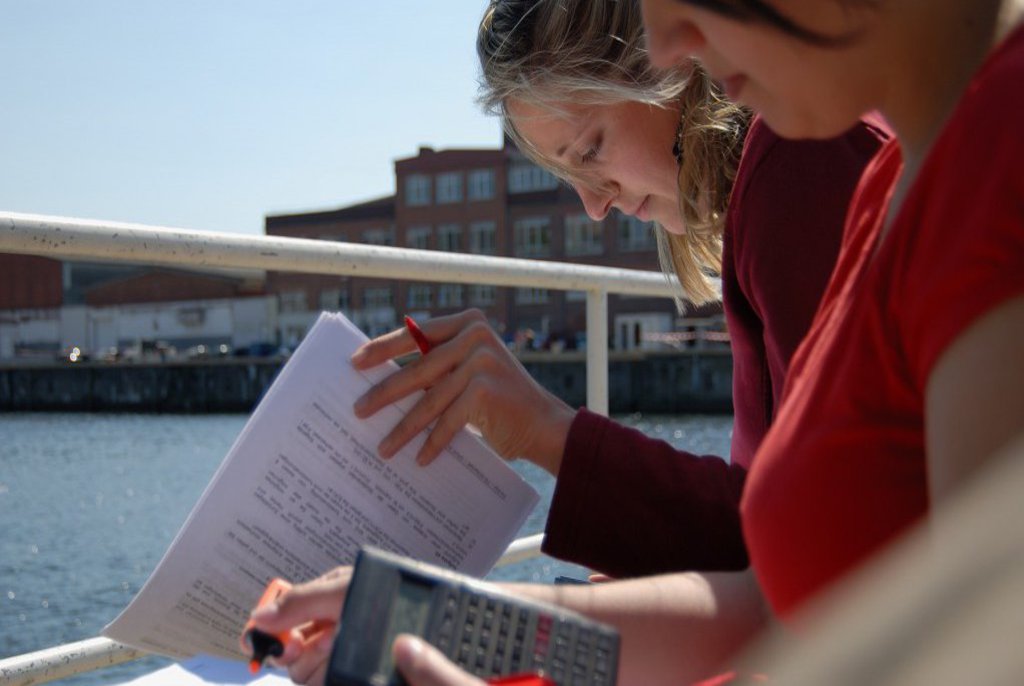 Zwei Frauen sitzen mit Schreibern und Rechnern ausgestattet am Wasser und arbeiten.