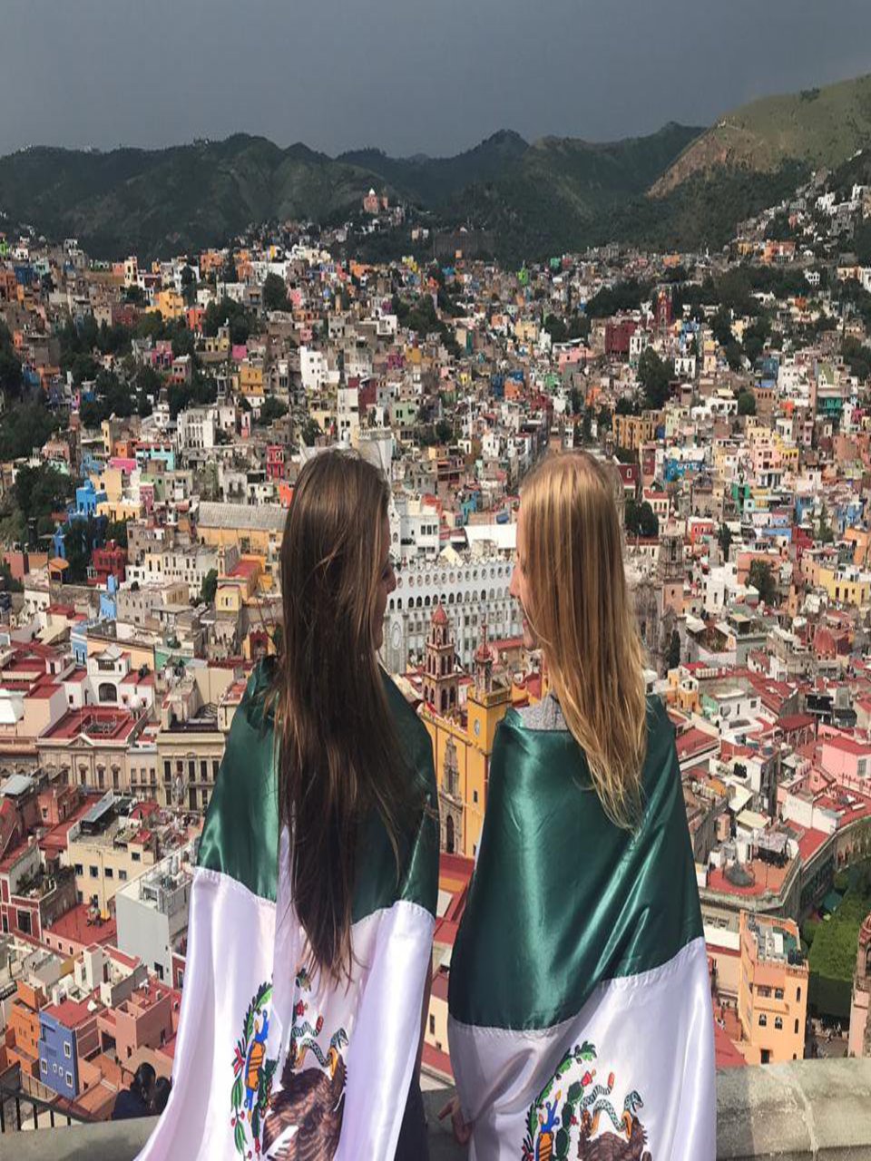Zwei Frauen posieren an der Brüstung eines Aussichtspunkts, von dem aus die Dächer einer Stadt zu sehen sind. Die Frauen haben sich die Flagge von Mexiko über die Schultern gelegt.
