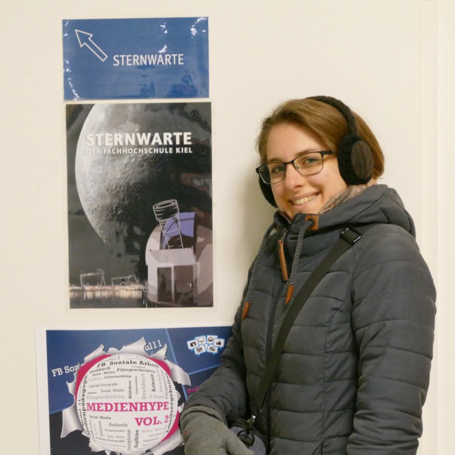 Eine Frau mit Winterjacke und Ohrenwärmern posiert vor Plakaten der Sternenwarte Fachhochschule Kiel.