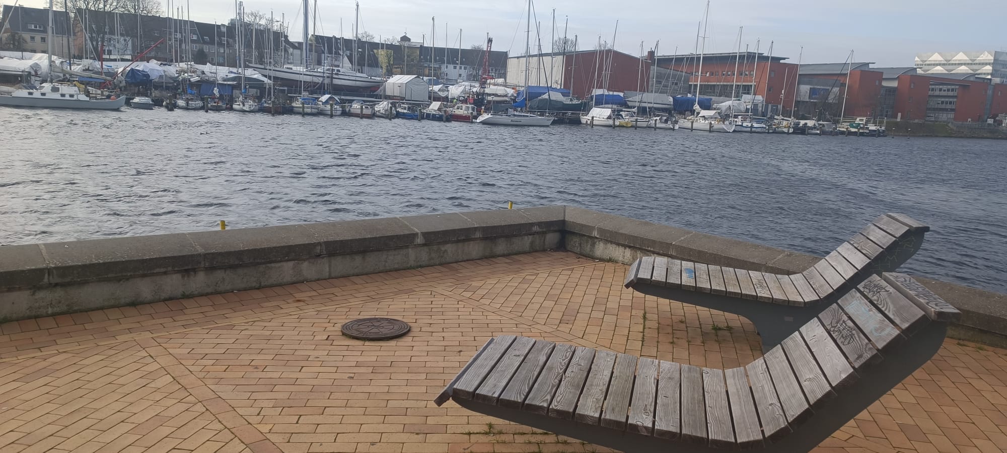 Auf dem Kai stehen zwei hölzerne Holzbänke, im Hintergrund sieht man Segelschiffe im Hafen liegen