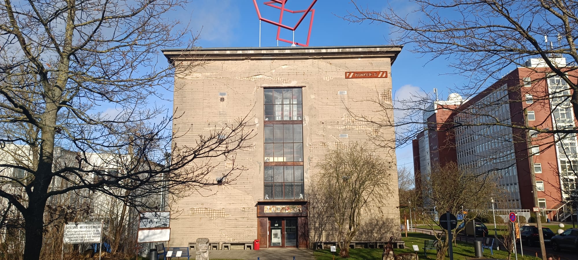 ein massiver Steinbau zwischen einem Baum und einem Backsteingebäude.