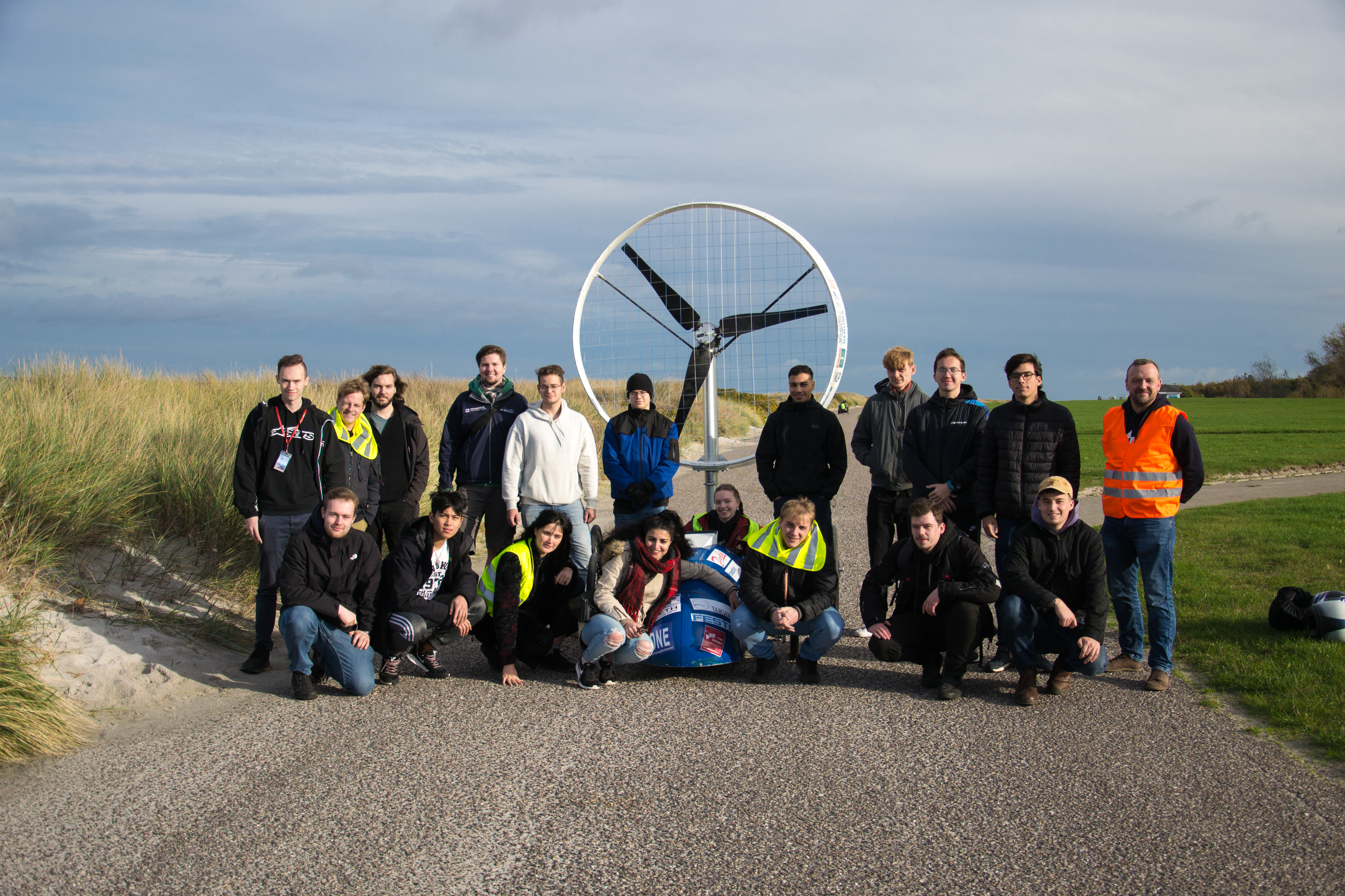 19 Menschen posieren für ein Gruppenbild. Im Hintergrund erkennt man Dünen und flaches Land.