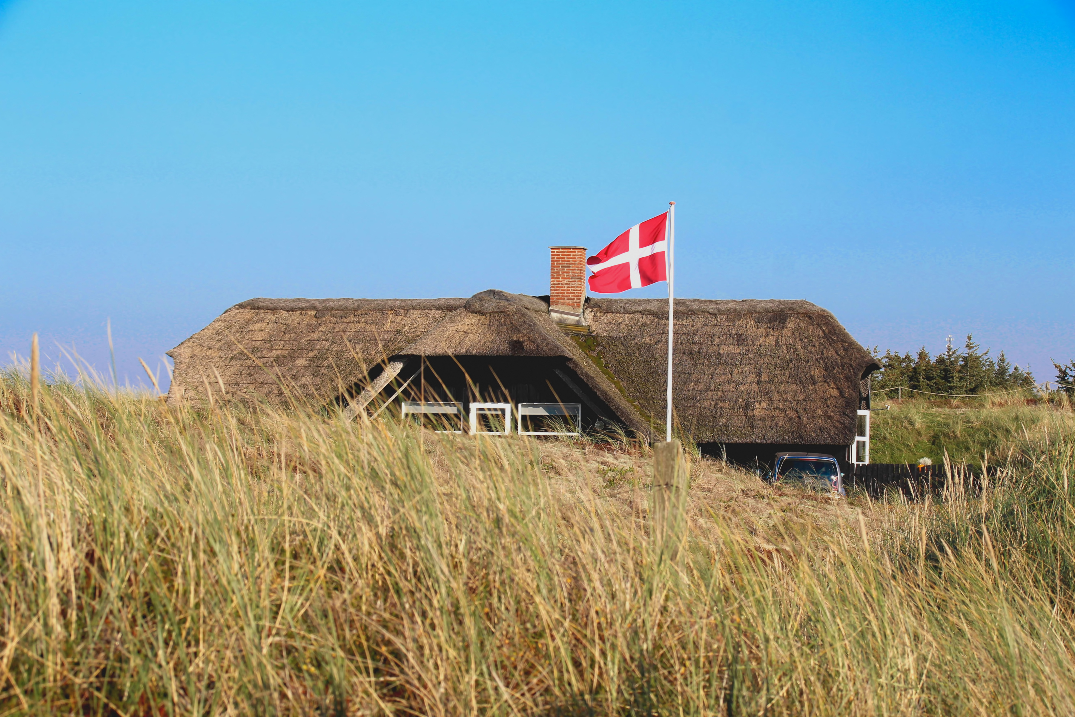 ein Reetdachhaus hinter Dünen, davor weht die Nationalflagge Dänemarks