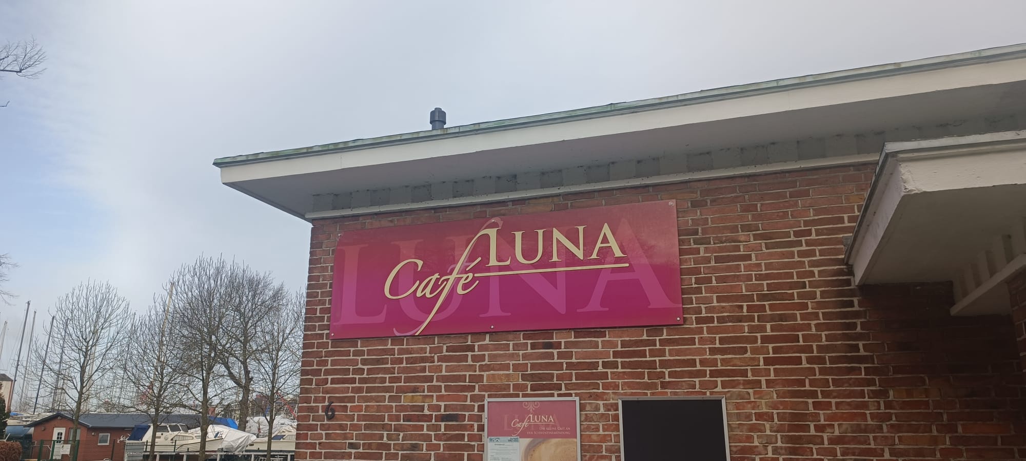 Das rote Ladenschild vom Café Luna hängt an der Hausecke. Im Hintergrund sieht man grauen Himmel.