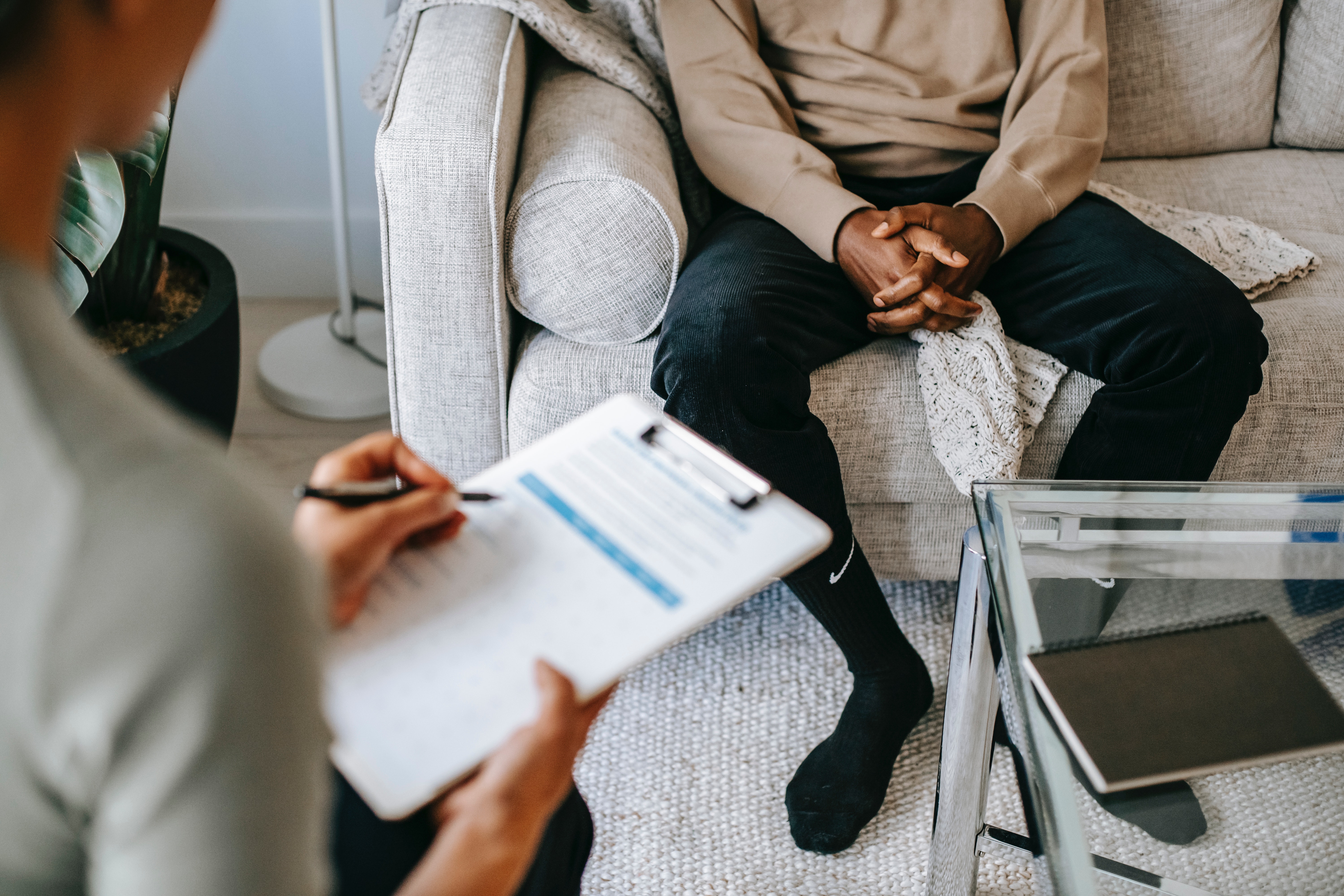 Man sieht zwei Personen im Therapie-Kontext. Die eine Person sitzt auf einem Sofa und hat die Hände im Schoß gefaltet, die andere macht Notizen auf einem Klemmbrett.