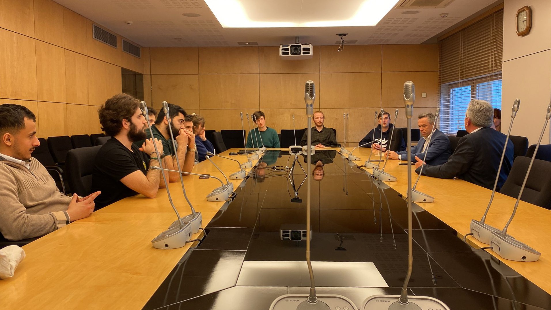 Menschen an einem großen Konferenzstisch, auf dem Tisch stehen Mikrofone