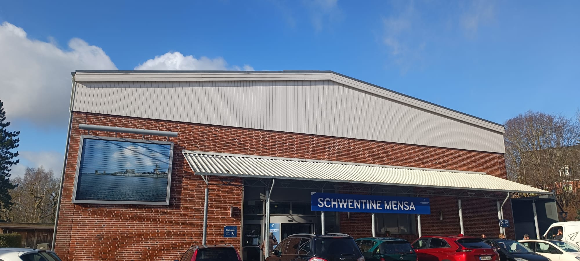 Ein Backsteingebäude mit der Aufschrift "Schwentine-Mensa". Im Vordergrund parken Autos, im Hintergrund ein beinahe wolkenloser Himmel.