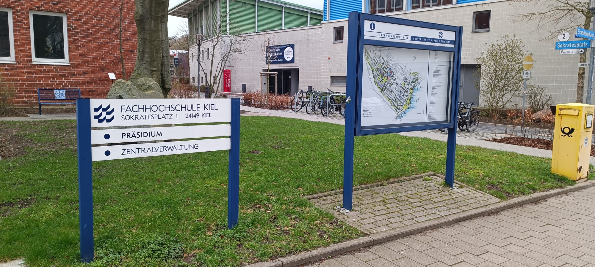 Im Bild sieht man im Vordergrund zwei Schilder. Zur Rechten den Lageplan des Campus, zur Linken das Ausweiseschild des Gebäudes, das im Hintergrund zu sehen ist.