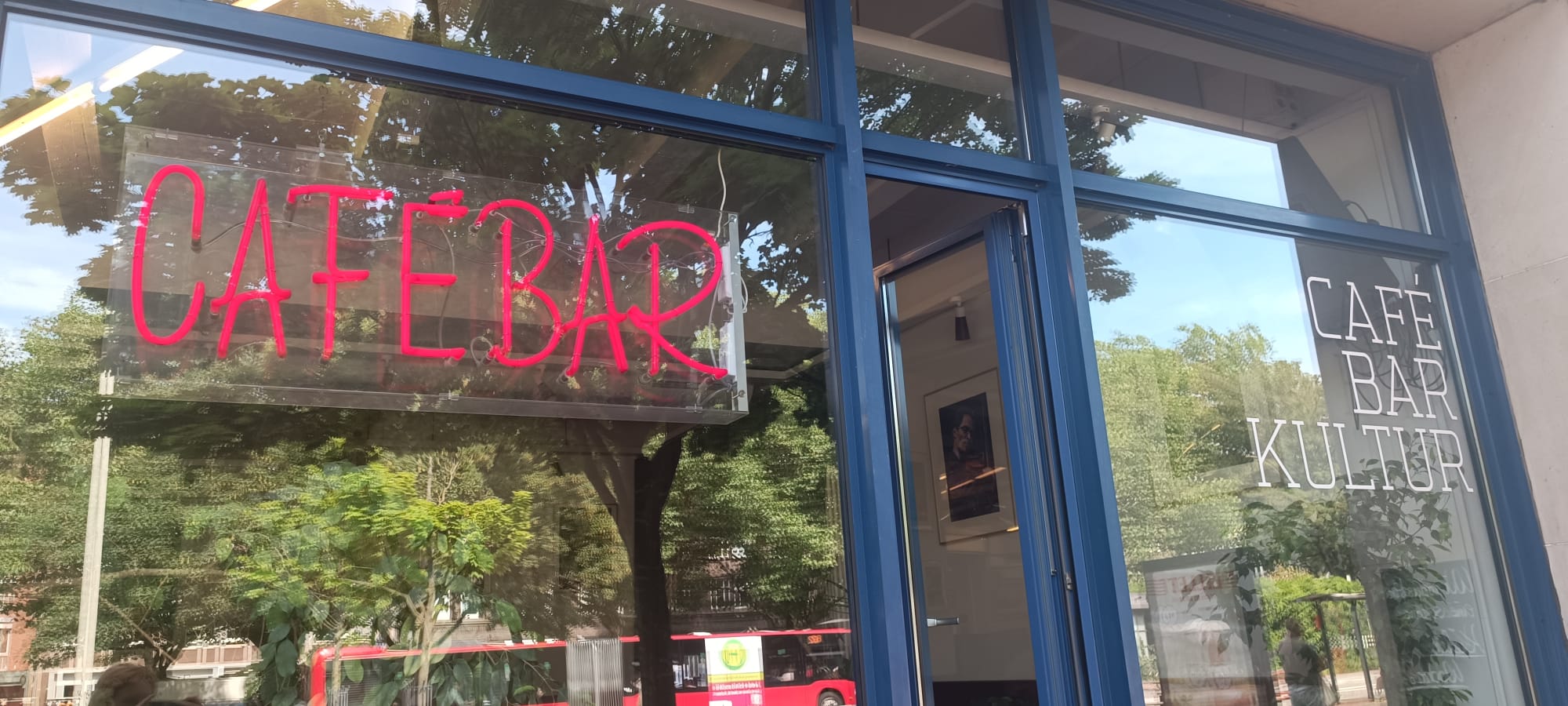 Ein Fenster, in welchem sich Bäume spiegeln. Eine Anzeige aus Neon-Röhren schreibt "Cafébar" auf die Scheibe.