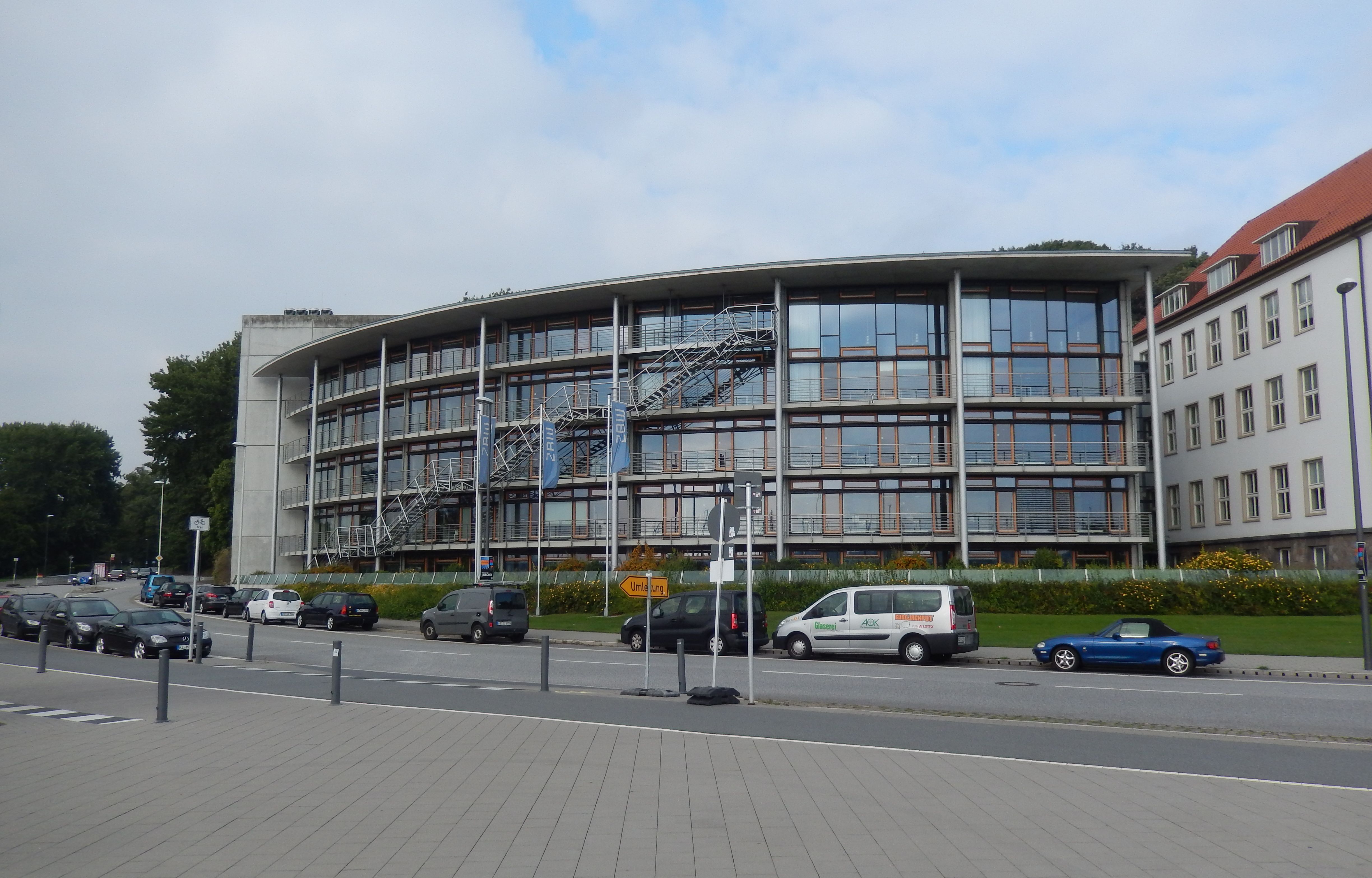 Man sieht ein rundes Gebäude mit Fensterfront. Im Vordergrund parken Autos.