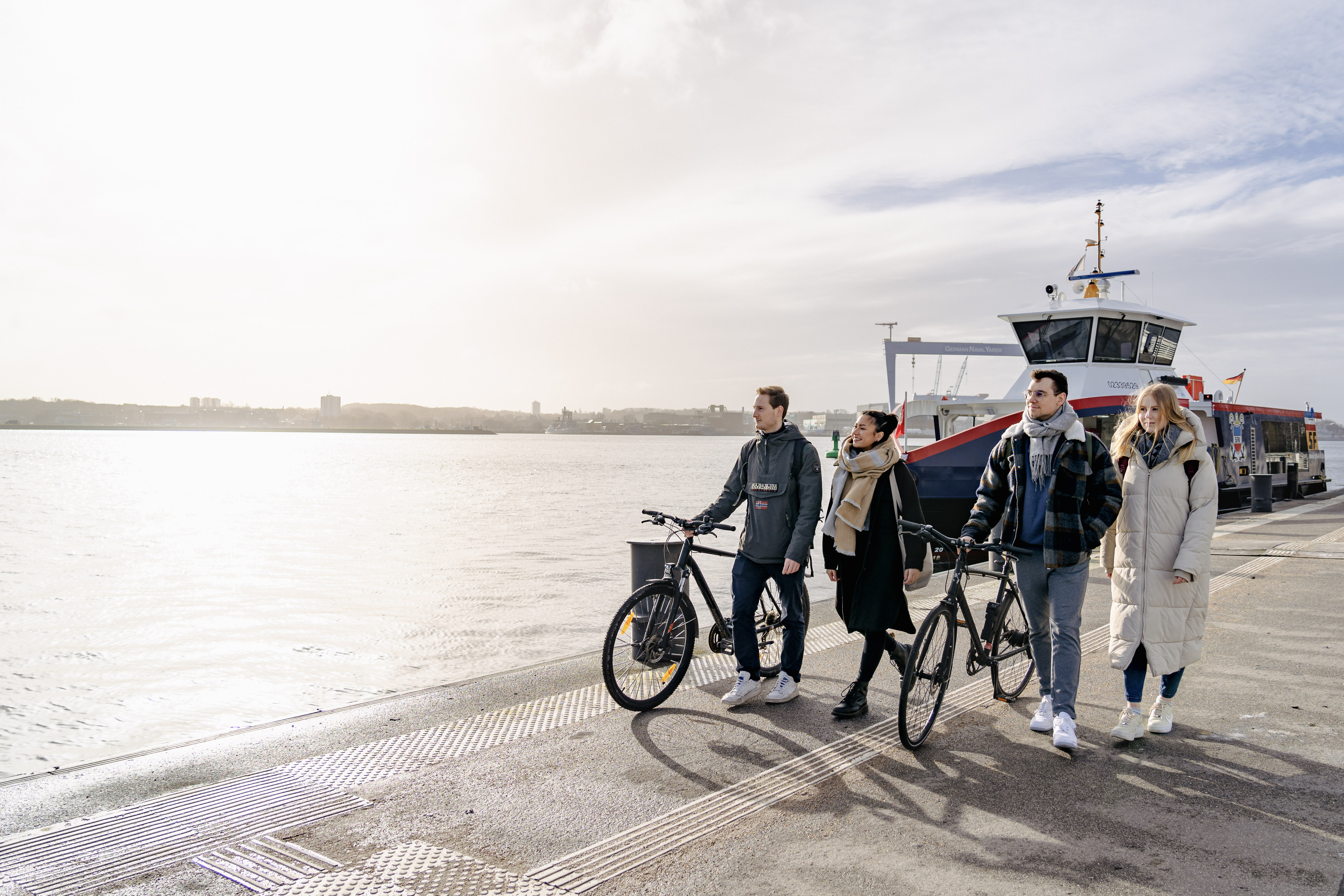 eine Gruppe von vier Personen geht entlang der Kiellinie im Sonnenschein, im Hintergrund ist der HDW-Kran zu sehen und eine Elektrofähre