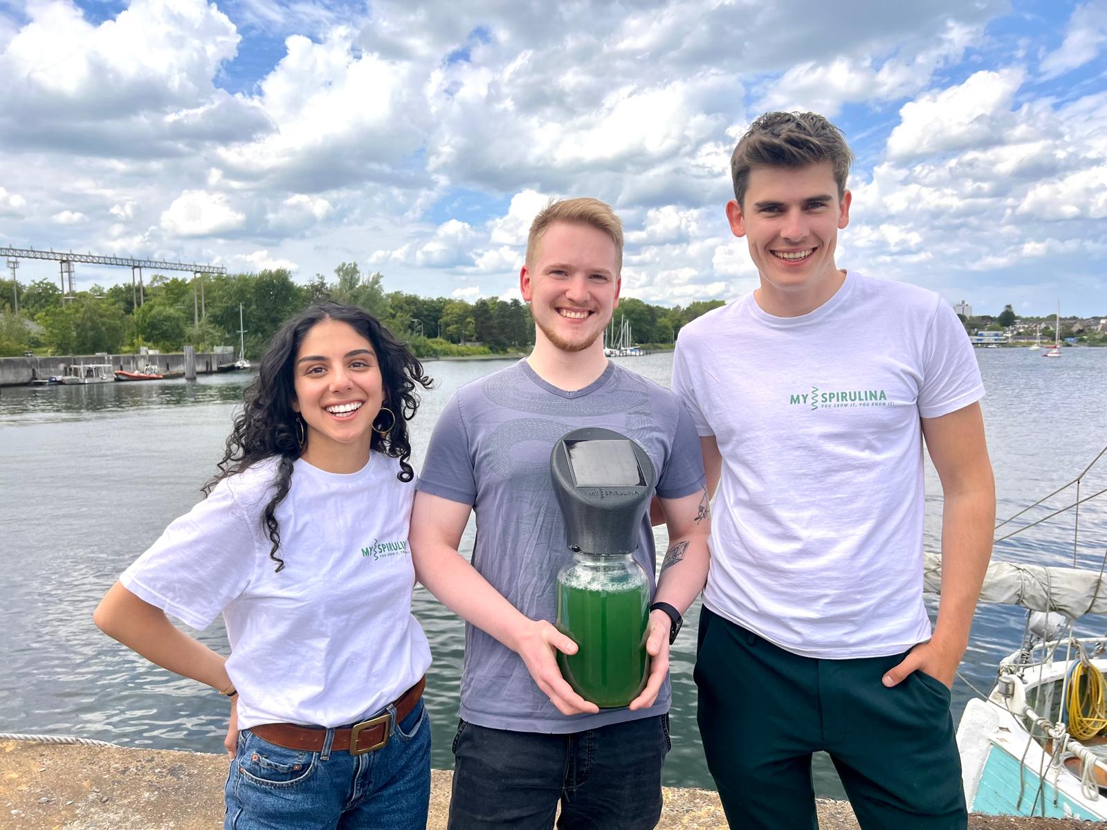 Die Gründer Sara Farzaneh Nejad, Jan Thode und Ben Schwedhelm stehen mit einem Glas Spirulina in der Hand am Waser..