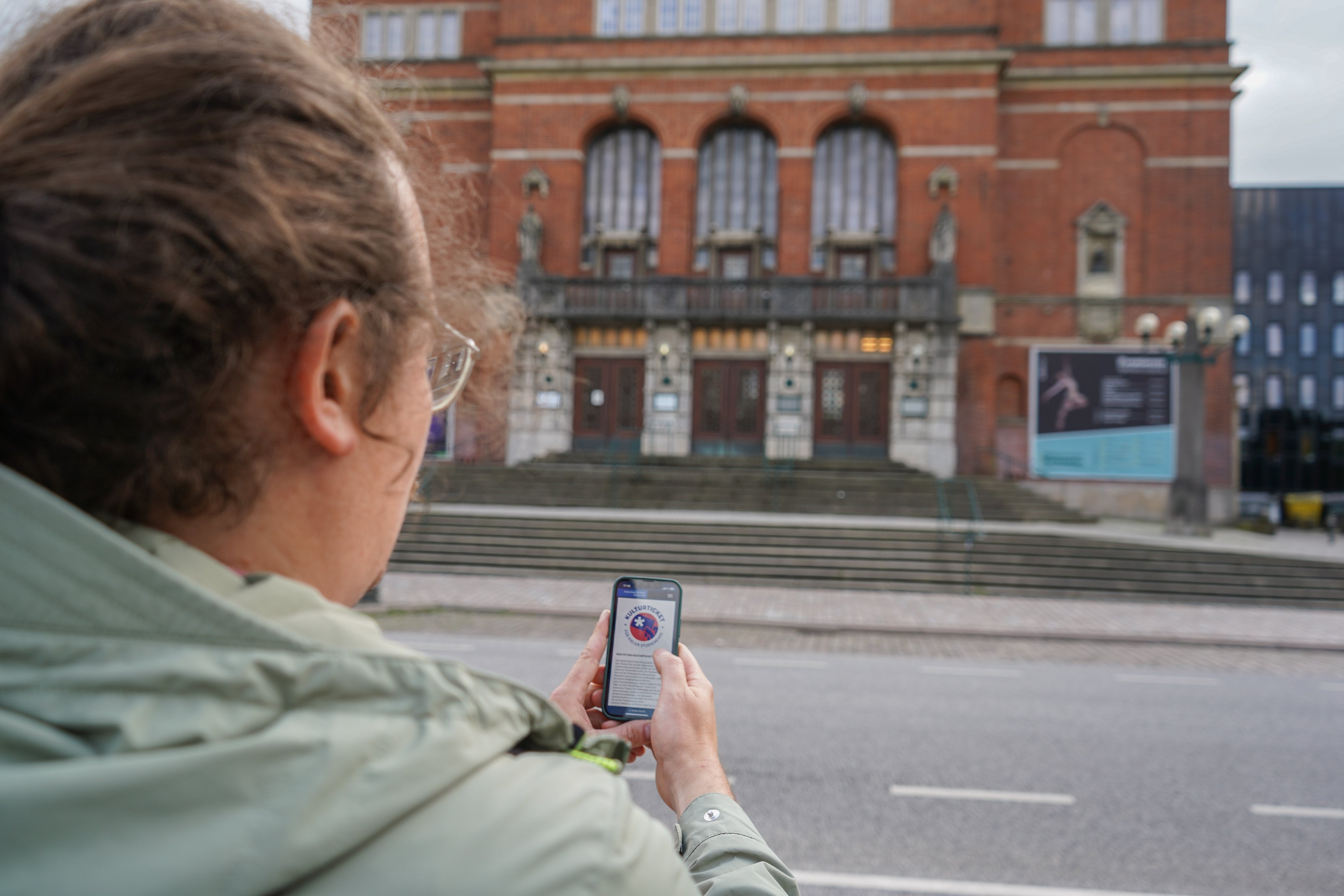 Ein Mann steht mit Handy in der Hand vor dem Kieler Opernhaus. Auf dem Handy sieht man die Website des Theaters, über die Studierendentickets gebucht werden können.