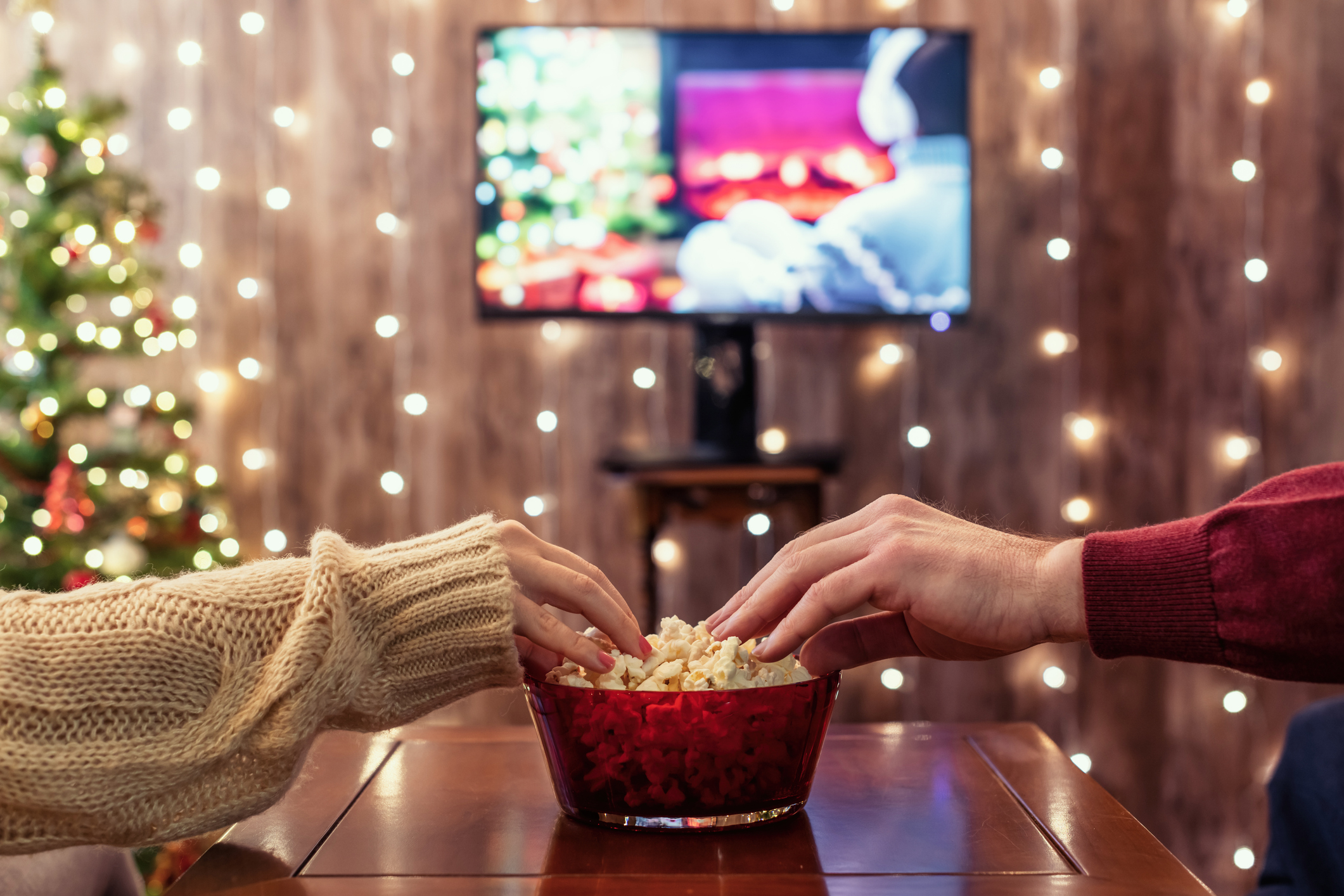 zwei Hände greifen in eine Popcornschale, dahinter ein Fernseher, drumherum weihnachtliche Dekoration