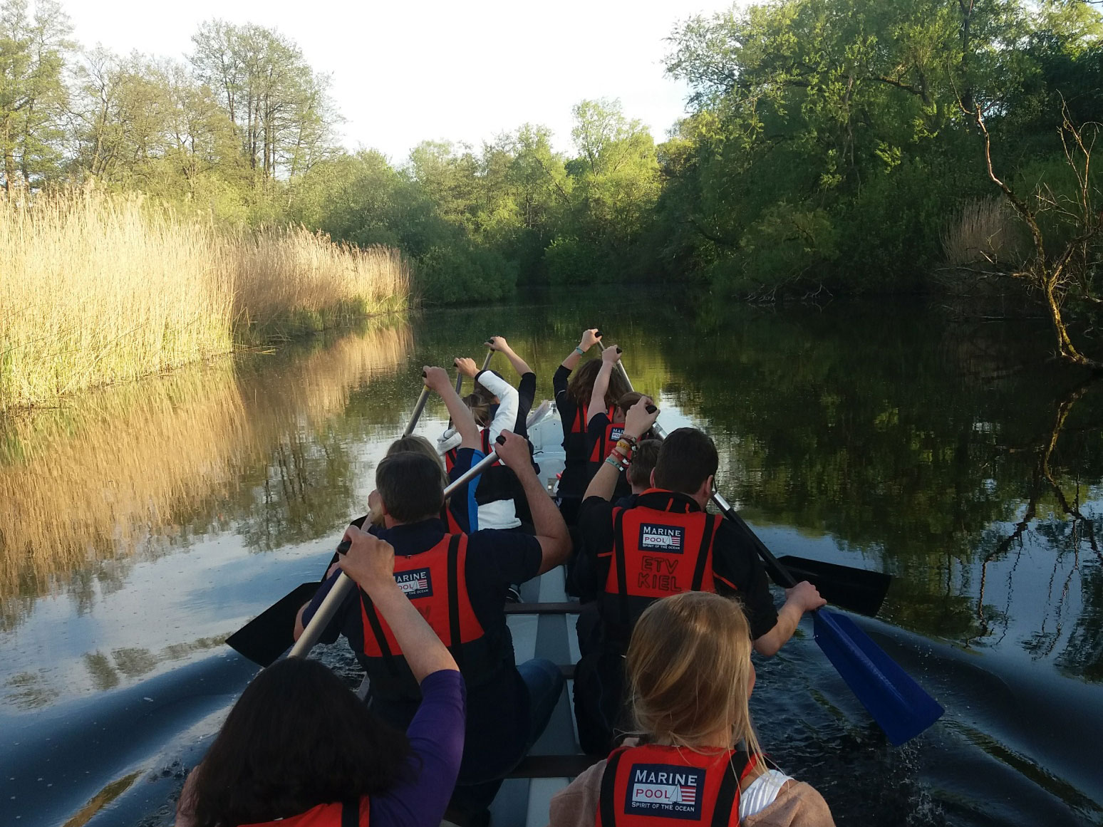 Eine Gruppe Studierender paddelt in einem Kanu einen Fluss entlang, alle heben Ihr paddel um zu nächsten Zug auszuholen.