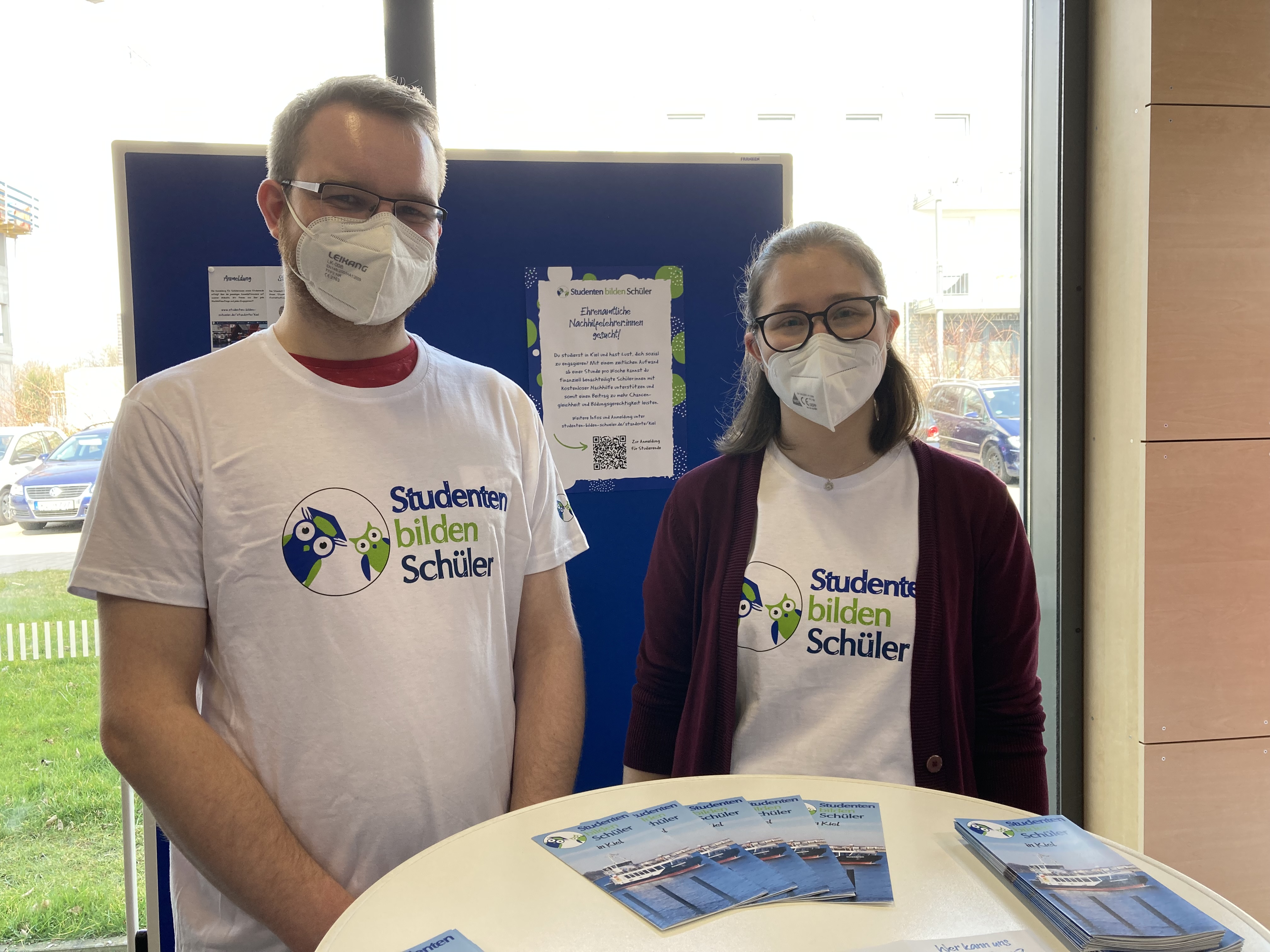 Ein junger Mann und eine junge Frau vor einer Infotafel. Sie tragen T-Shirts mit dem Aufdruck "Studenten bilden Schüler"