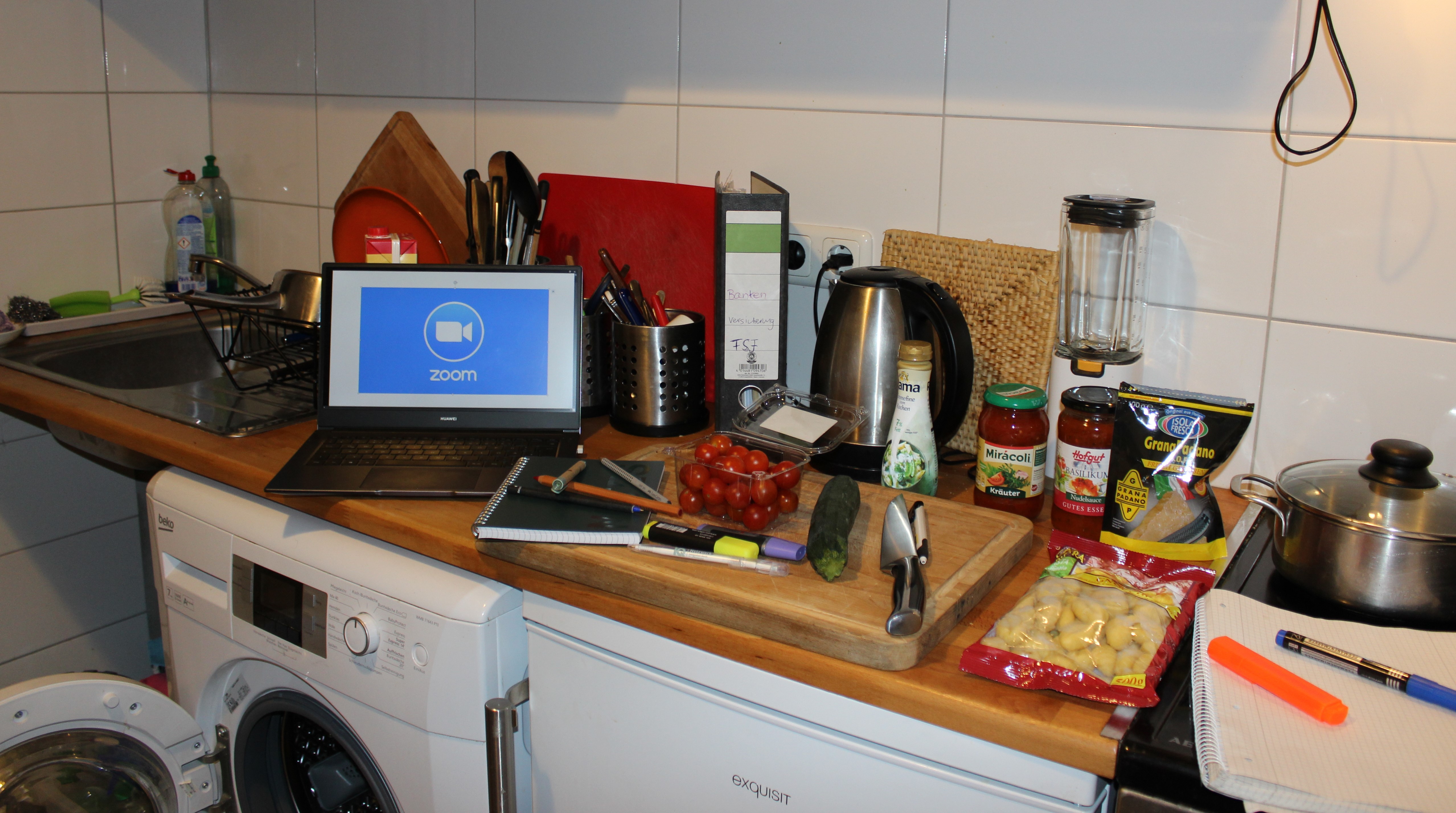 Laptop in der Küche auf der Arbeitsfläche, auf dem Display ist der Videokonferenz-Dienst Zoom geöffnet, auf und neben dem Schneidebrett liegen Lern- und Kochutensilien