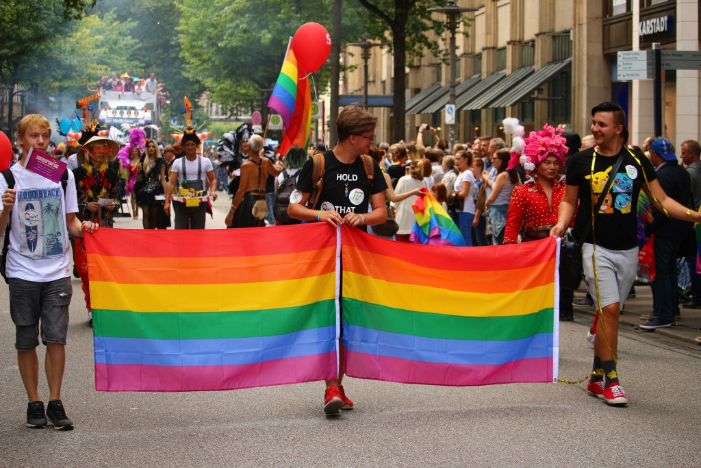 Drei Männer halten zwei Regenbogenflaggen und marschieren mit einer Menschenmenge.