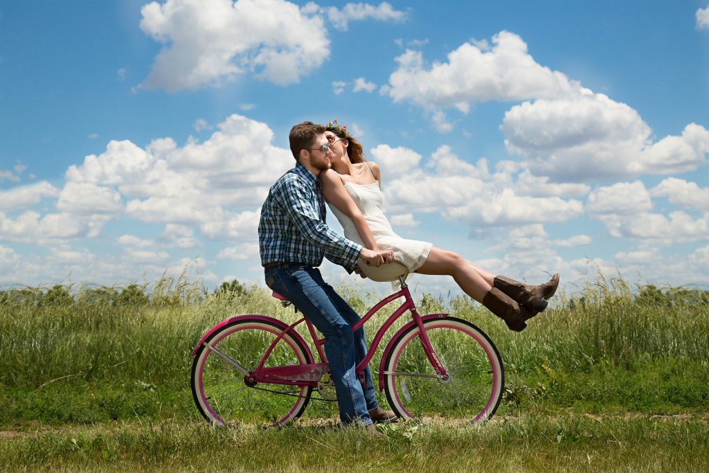 Ein Mann sitz auf einem fliederfarbenen Fahrrad auf einer Landstraße. Auf dem Lenker sitzt eine Frau und wendet den Kopf um dem Mann einen Kuss zu geben.
