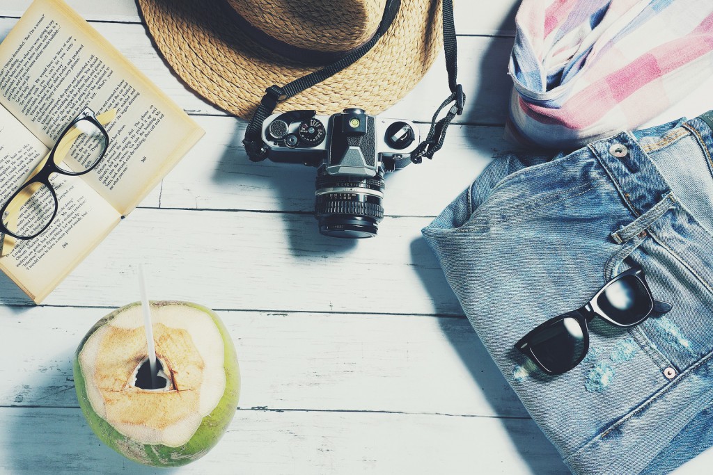 Eine Kokosnuss, ein Buch mit Brille darauf, ein Hut, eine Kamera, ein Schal und eine Jeans mit Sonnenbrille darauf liegen auf einem Holztisch.