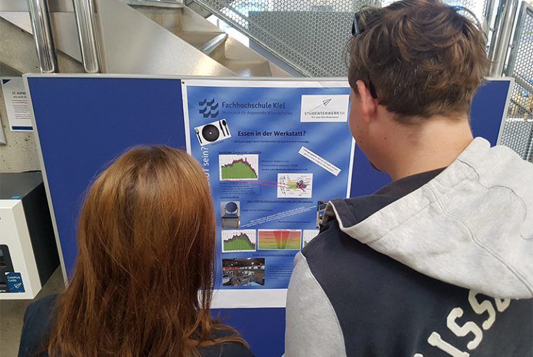 Eine Frau und ein Mann stehen mit dem Rücken zur Kamera und lesen ein Plakat.