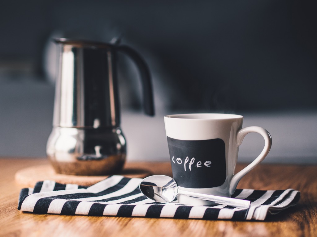 Eine Kaffeetasse steht auf einem blau weisen Tuch, im Hintergrund steht eine Espressokocher.